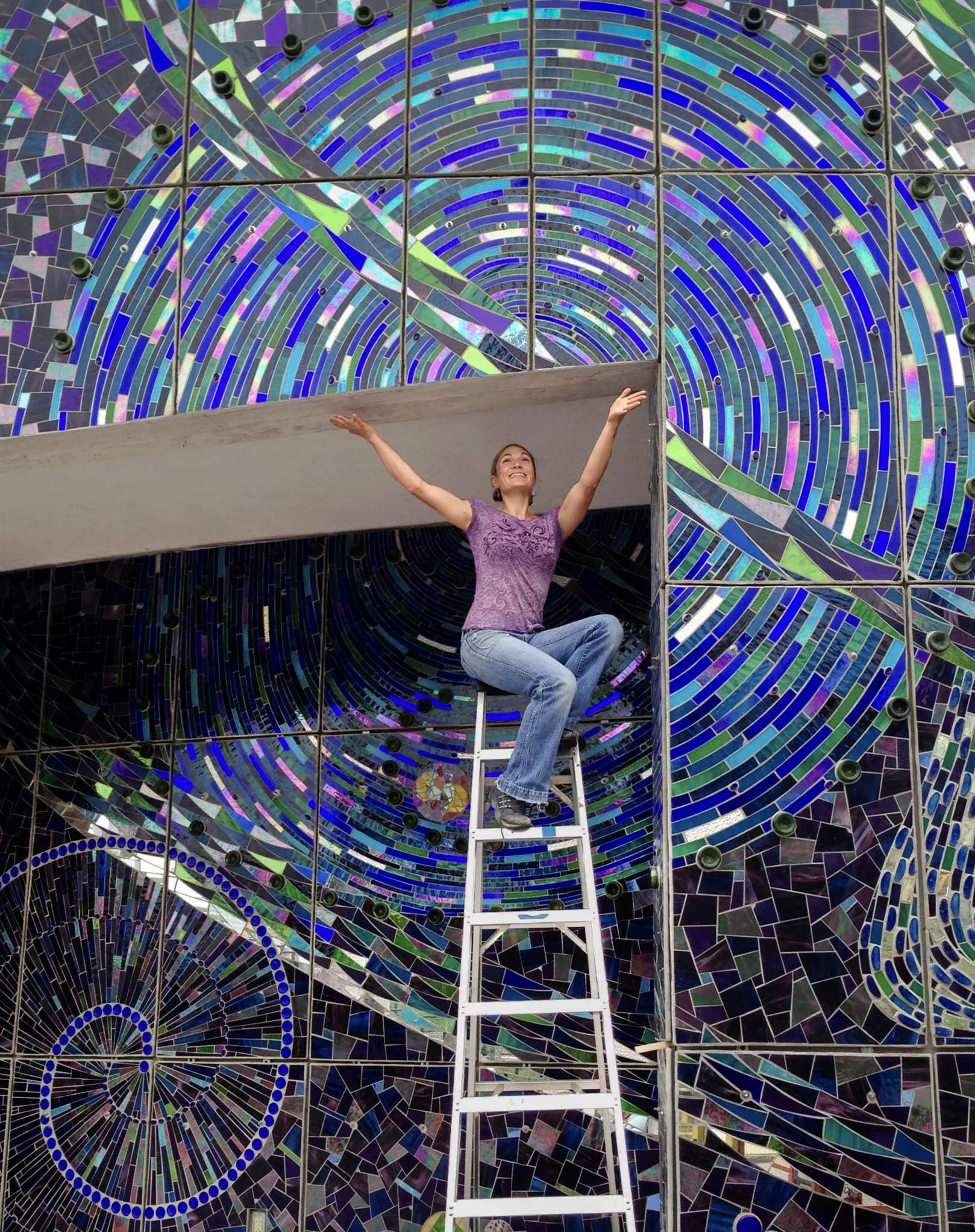 Mari Gardner on a ladder, arms raised, in front of her completed mosaic Aurora Borealis